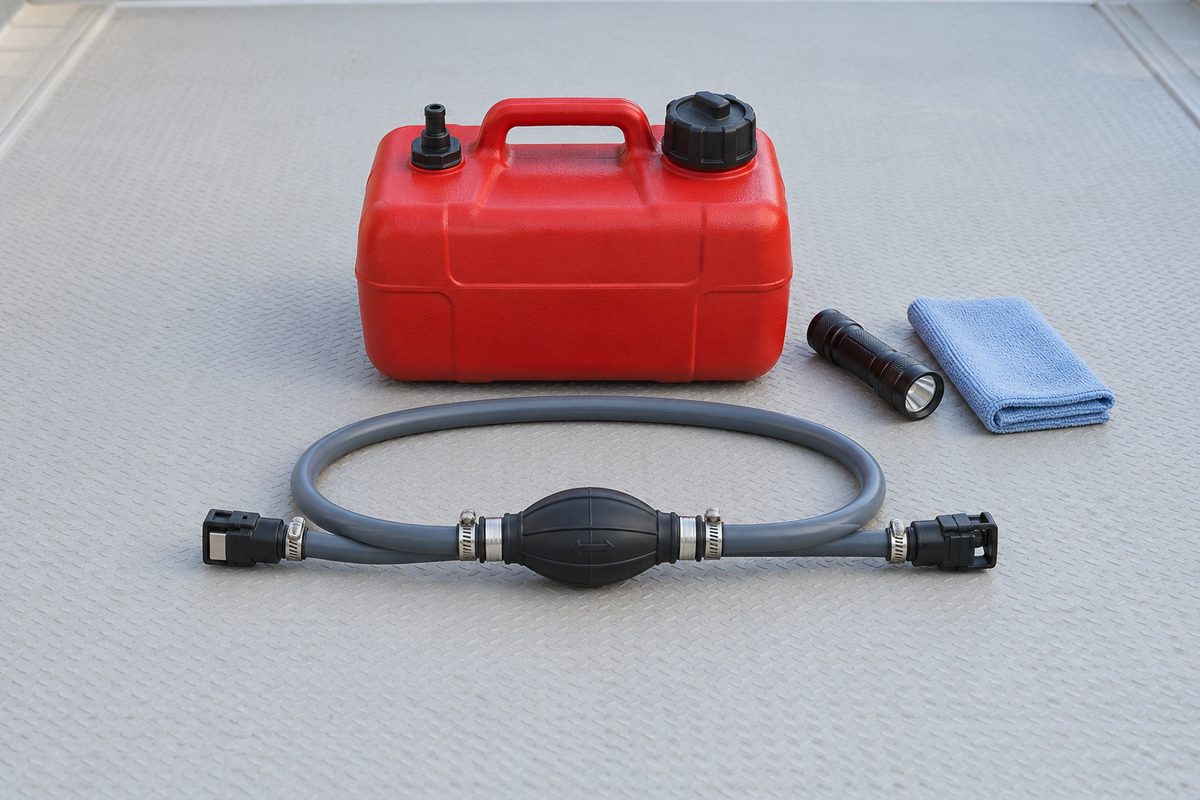 A gray outboard fuel line with a black primer bulb, hose clamps, and quick-connect fittings in front of a red portable fuel tank, beside a flashlight and blue microfiber towel on a gray deck.